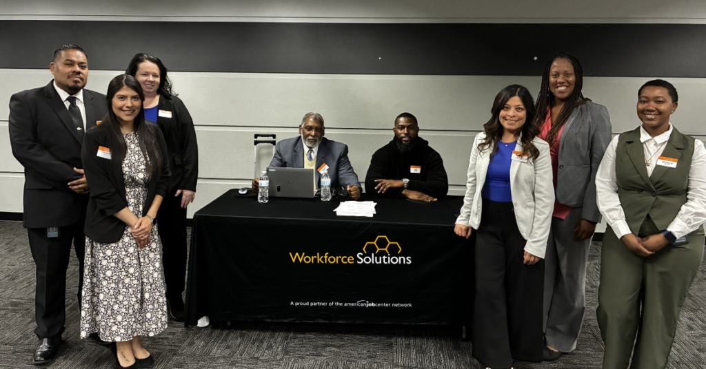A group of seven Workforce Solutions Staff, five standing and two seated at a table with laptops. Smiling, wearing business attire. Workforce Solutions logo visible.