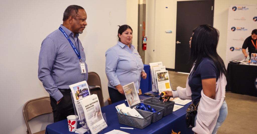 Two people stand behind a table at a job fair, talking to a visitor. The table has brochures, pens, and a laptop, creating a welcoming and informative atmosphere.