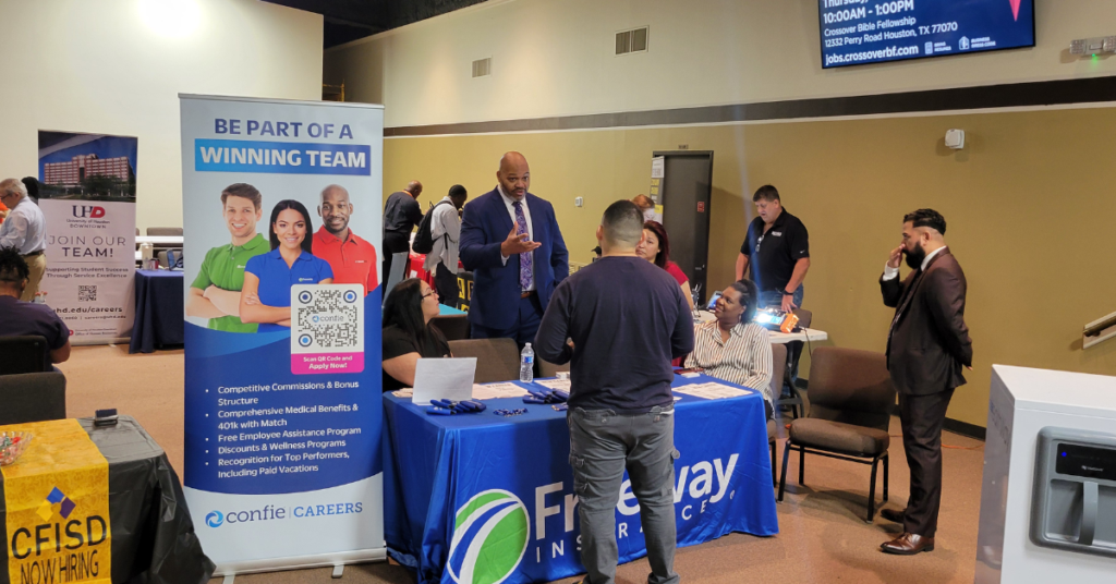 A career fair scene with multiple booths. Professionals engage at a table with a "Freeway Insurance" banner. The atmosphere is busy and interactive.