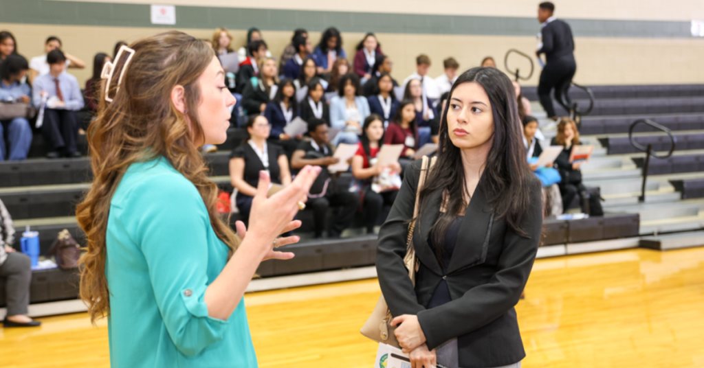 Two women in conversation at a busy event in a gymnasium. One gestures expressively, while the other listens intently. Background shows people seated.
