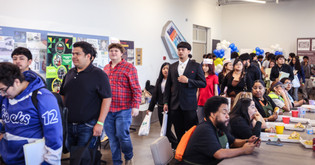 A diverse group of people walks through a bright room, wearing casual and formal attire. Some sit at tables with plates, creating a lively atmosphere.
