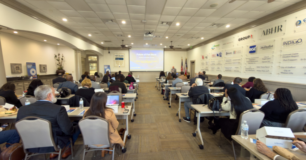 A conference room with people seated at tables facing a projected presentation. Attendees are engaged, suggesting a professional, focused atmosphere.