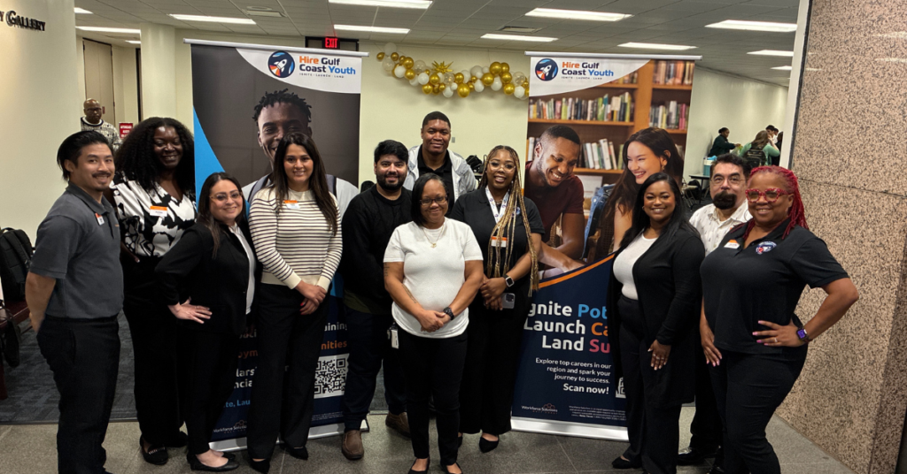 A diverse group of eleven people stands smiling in an office lobby. Behind them are two banners promoting "Hire Gulf Coast Youth" with vibrant images and text.