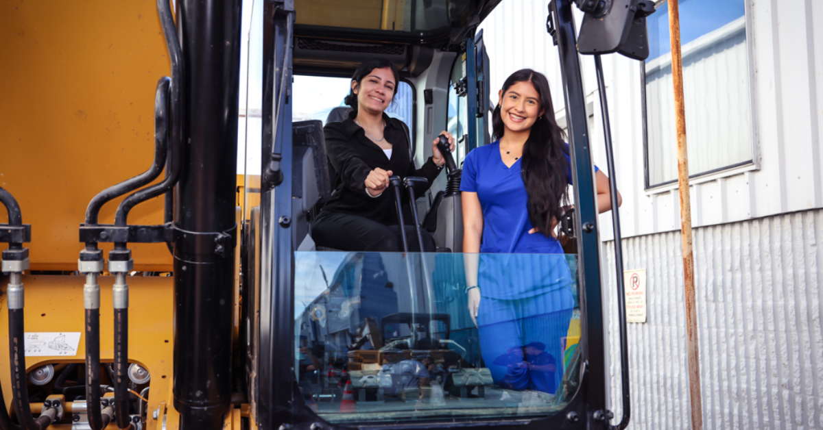 ARP_MC3 Apprenticeship Readiness Program A woman in a black shirt sits in the cab of a yellow excavator while another woman in blue scrubs stands beside the open door of the machinery.