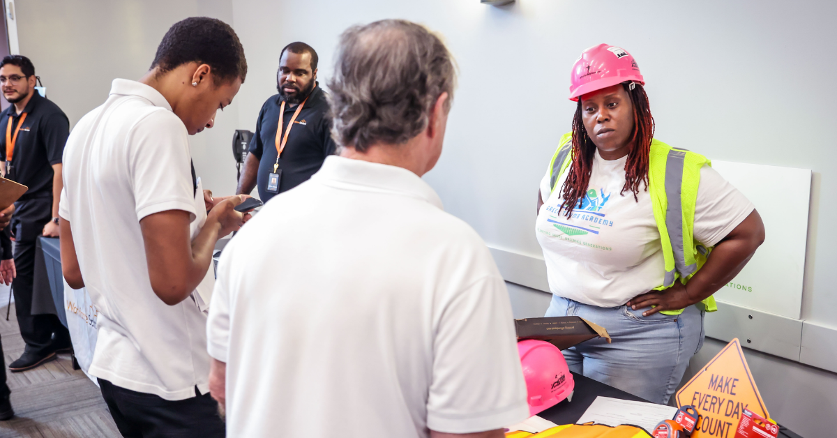 ARP_MC3 Apprenticeship Readiness Program A woman wearing a pink hard hat and high-visibility vest speaks with people at a career fair booth with a sign that says Make Every Day Count.