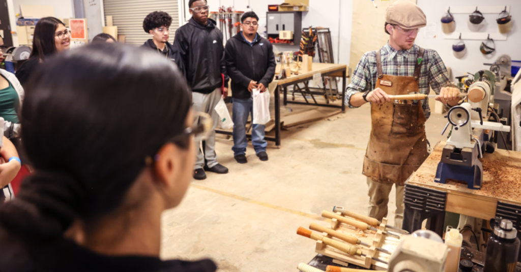 A woodworking instructor wearing a leather apron and cap demonstrates woodturning on a lathe for a group of students in a bright workshop setting.