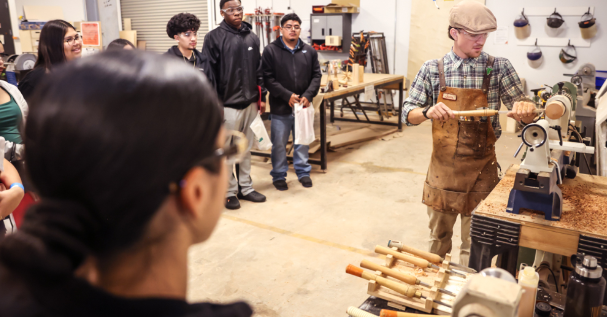 A woodworking instructor wearing a leather apron and cap demonstrates woodturning on a lathe for a group of students in a bright workshop setting.