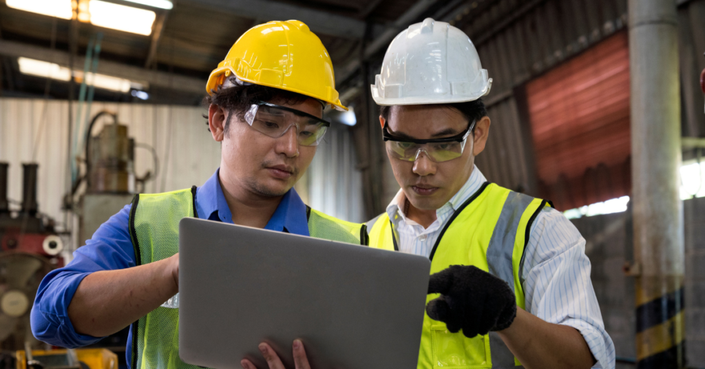 Two factory workers in safety gear, including helmets and vests, focus intently on a laptop screen in an industrial setting, collaborating on a task.