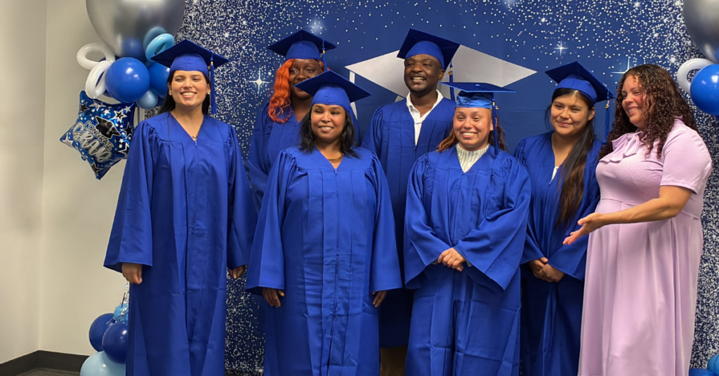 A group of graduates wearing blue caps and gowns stands smiling against a sparkling backdrop.