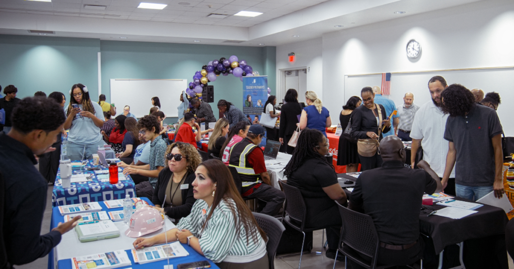 Be Successful Job Fair, with diverse attendees and recruiters at tables covered with brochures. The room is lively, with conversations and a balloon arch in the background.