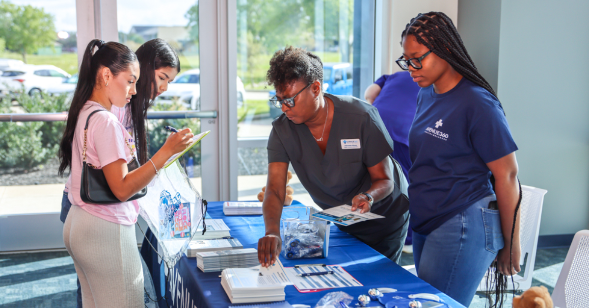 Four women engage at an informational booth with pamphlets and pens. One woman signs a form, while others observe.