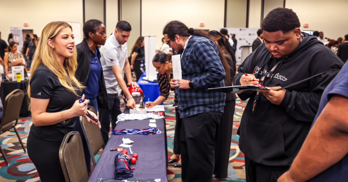 A job fair with diverse attendees at a booth, engaging with a woman handing out materials.