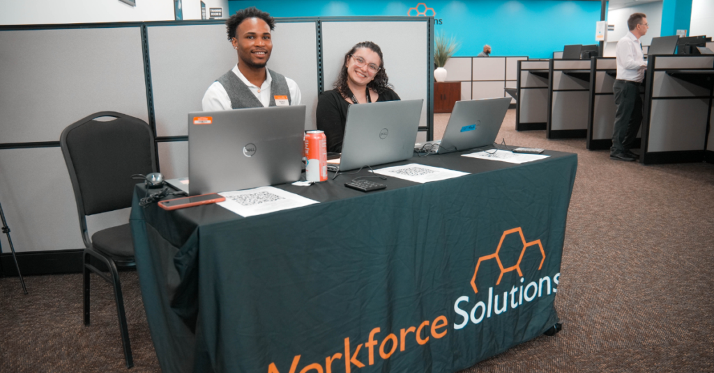 Two smiling individuals sit behind a table with laptops and a "Workforce Solutions" tablecloth. The setting is an office environment with cubicles.