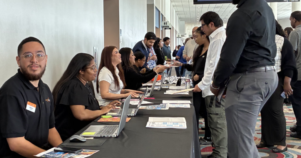 A busy job fair scene with diverse attendees lined up at tables. Staff seated behind laptops help visitors with inquiries, creating a collaborative atmosphere.