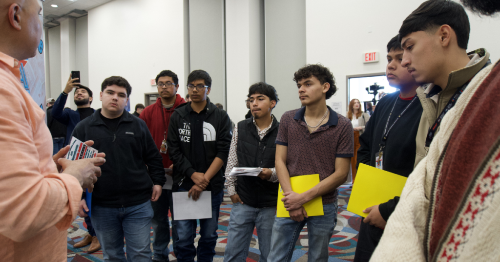 A group of young men attentively listens to an employer at a job fair. They hold notebooks and folders, showing engagement and focus.