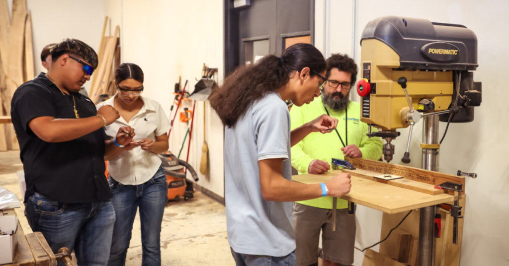 Instructor guides students using woodworking equipment during a hands-on skilled trades training session.