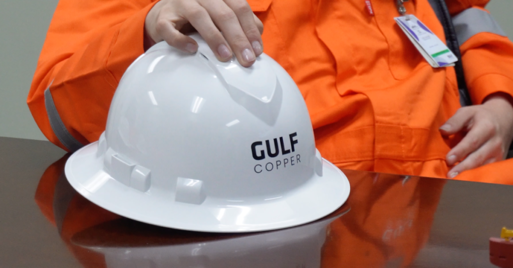 Worker in orange protective workwear rests a hand on a white Gulf Copper hard hat, representing skilled trades and industrial career opportunities.