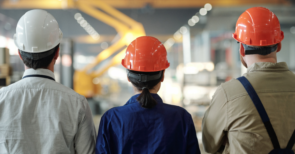Three manufacturing professionals wearing safety helmets stand inside an industrial facility, looking toward production equipment and machinery on the factory floor.