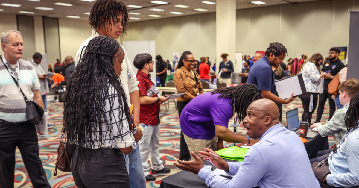 A job seeker speaks with an employer seated at a recruitment table during a crowded hiring event, while other attendees meet with recruiters and explore employment opportunities throughout the venue.