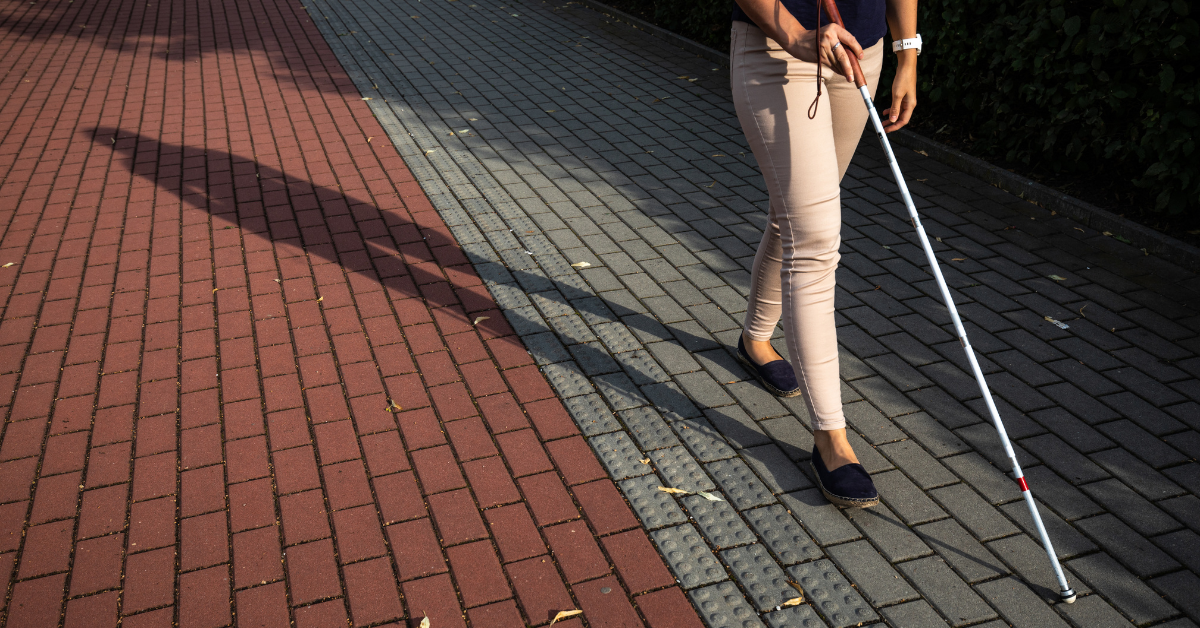 person with a visual impairment uses a white mobility cane to navigate a paved pedestrian walkway with tactile paving, demonstrating accessible mobility and independent travel.