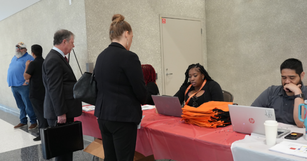 Job seekers speak with event representatives at a hiring event registration table, where staff members assist attendees using laptops and provide information about employment opportunities.