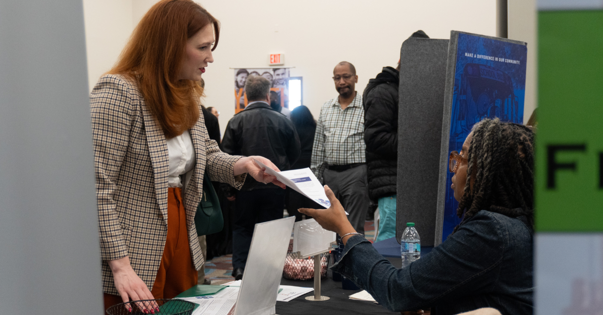 A job seeker in a plaid blazer speaks with a seated recruiter and exchanges informational materials at an employment booth during a hiring event, with other attendees networking in the background.