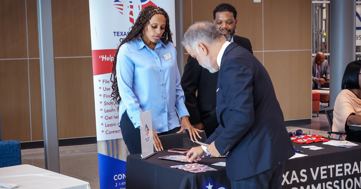 A man in a navy suit reviews informational materials at the Texas Veterans Commission booth while two representatives assist him during a veterans hiring event.