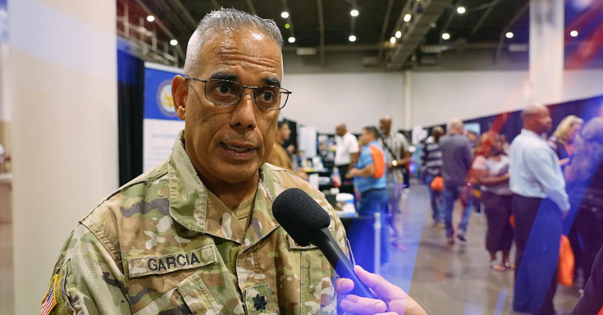 Military representative Garcia, wearing a camouflage uniform, speaks into a microphone during an interview at a workforce event, with attendees and informational booths visible in the background.