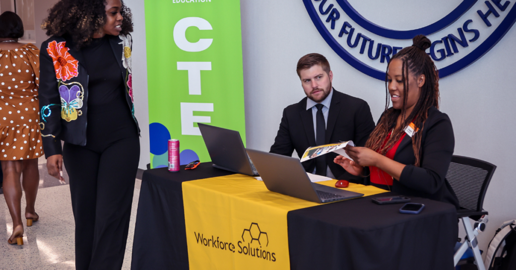 Two Workforce Solutions representatives seated behind a registration table with laptops review informational materials while speaking with an attendee at a career event. A Workforce Solutions banner and branded tablecloth are visible.