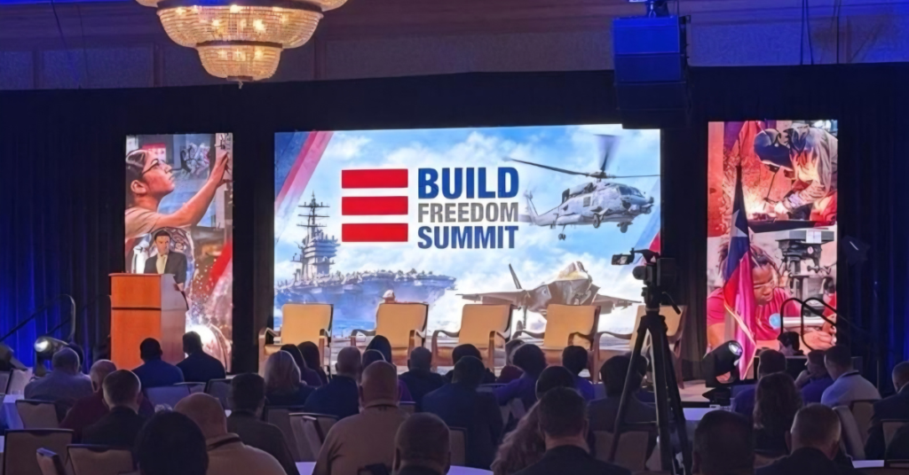 Attendees seated at the Build Freedom Summit watch a speaker at a podium, with a large stage display featuring military aircraft, a naval ship, and the event's logo.